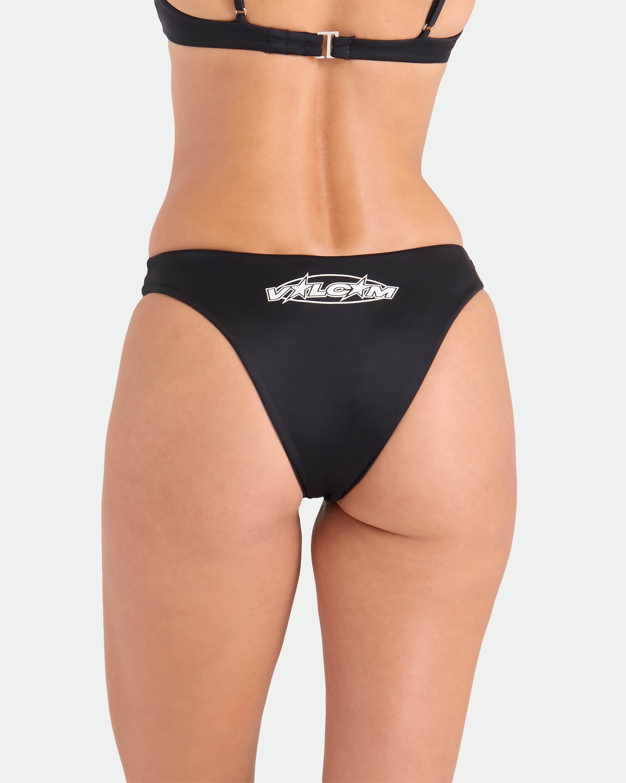 A woman models the Volcom Women's Coco Legacy Drop 90S V Bottom bikini—black, featuring a white “ALGZT” logo on the back. This high-leg style is made from recycled nylon for sustainability and is shown against a plain white background.