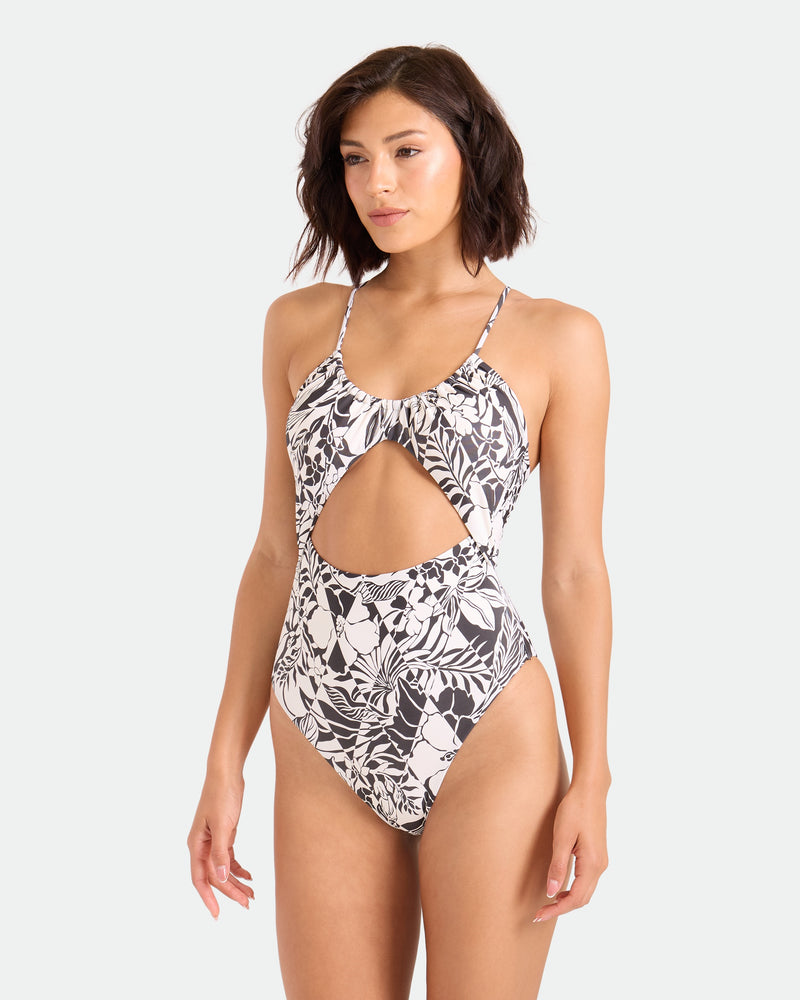 A woman wears the Volcom Women's Hibiscus Diamond Keyhole One-Piece, a black and white floral recycled-nylon swimsuit with a front cutout and thin straps, standing against a plain light background.
