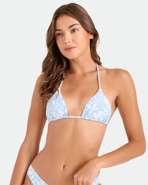 A woman with long brown hair models the Volcom Women's Tribal Stone Triangle Top—a light blue and white patterned bikini with adjustable tie straps—posing against a plain light background.
