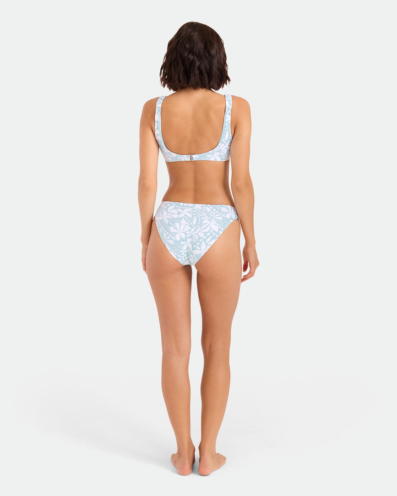 A woman stands barefoot facing away, wearing the Volcom Women's Tribal Stone Scoop Bralette bikini top in light blue and white floral recycled nylon. Her arms are relaxed at her sides, set against a plain white background.