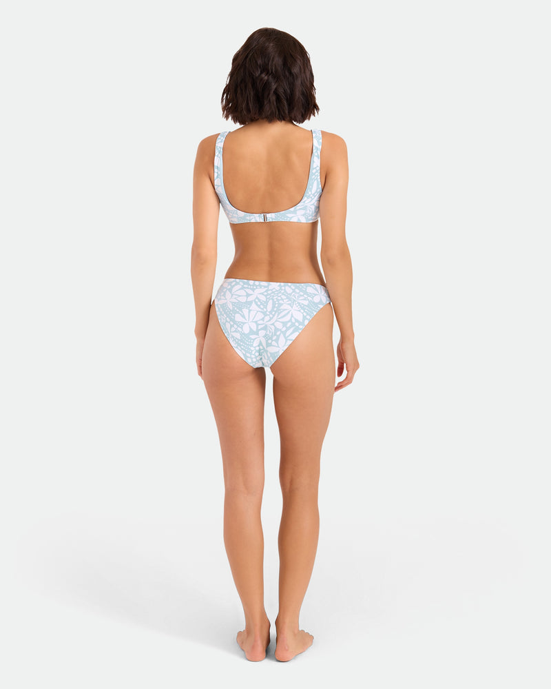 A woman stands barefoot with her back to the camera, wearing Volcom’s Women’s Tribal Stone Hipster Full Bikini Bottom in Summer Song, made from recycled nylon. The background is plain white.