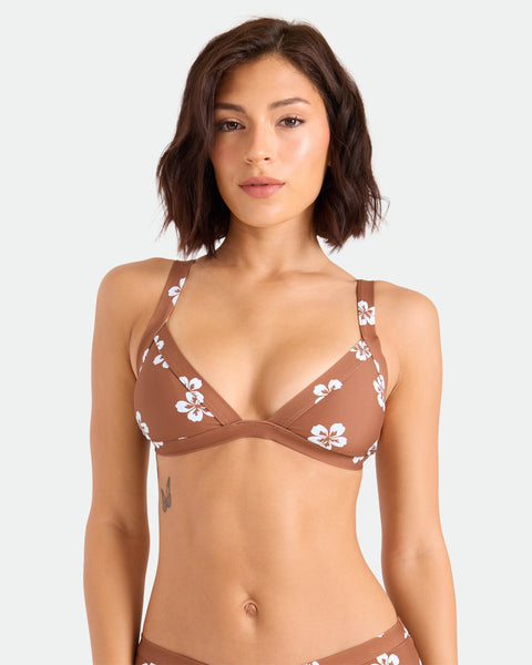 A woman with short brown hair wears the Volcom Women's Coco Retro Bloom Banded Triangle Top, a medium-support brown bikini with white floral patterns made from recycled nylon, standing against a plain light background.