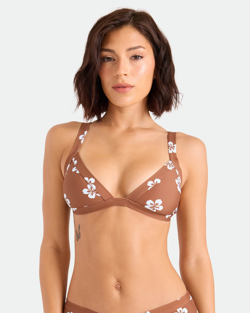 A woman with short brown hair wears the Volcom Women's Coco Retro Bloom Banded Triangle Top, a medium-support brown bikini with white floral patterns made from recycled nylon, standing against a plain light background.
