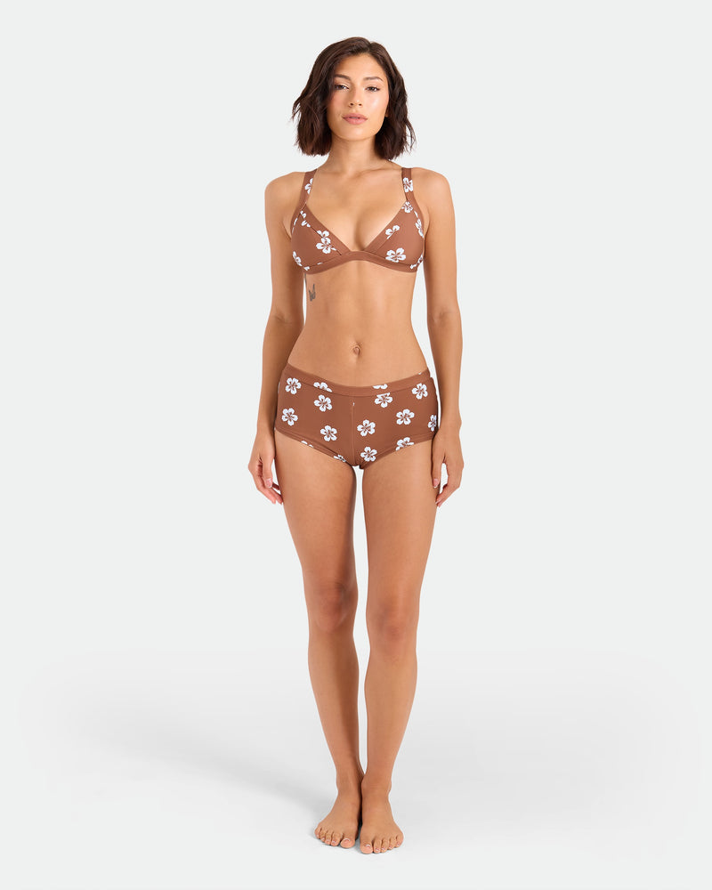 A woman with short brown hair stands barefoot against a plain background, wearing the Volcom Women's Coco Retro Bloom Banded Triangle Top—a brown bikini top with white floral patterns made from recycled nylon. Her arms are relaxed at her sides.