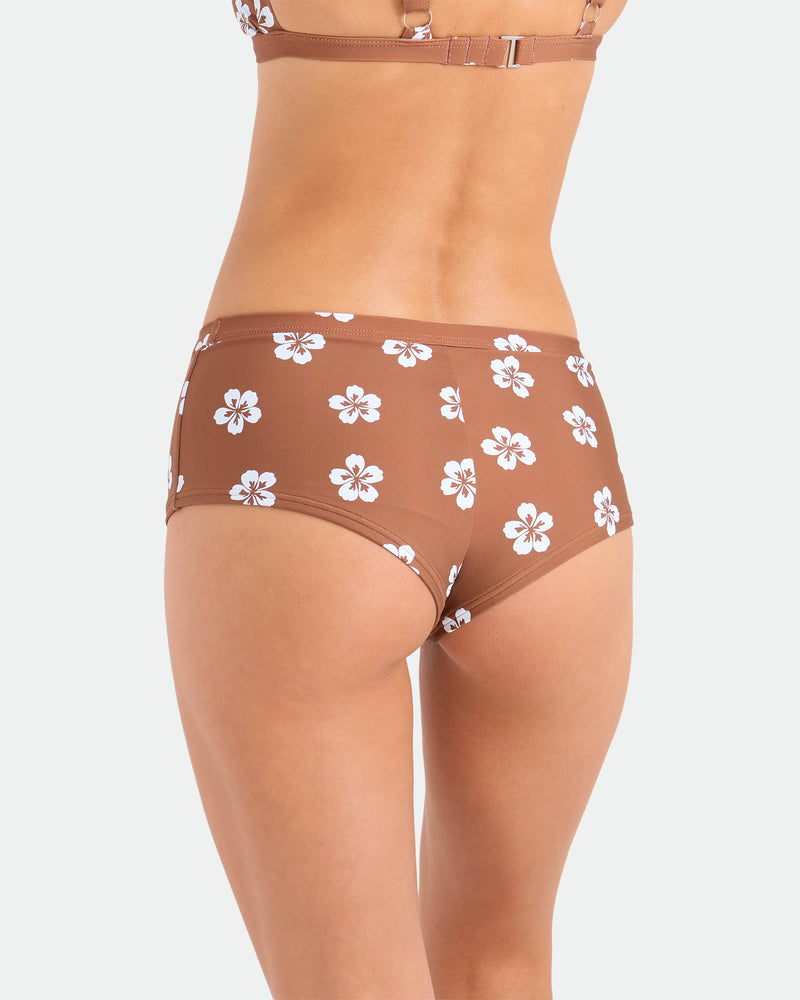 A woman is shown from the back wearing Volcom Women's Coco Retro Bloom Surf Shorts in brown with a white floral print and cheeky coverage, paired with a matching recycled nylon bikini top, against a plain white background.