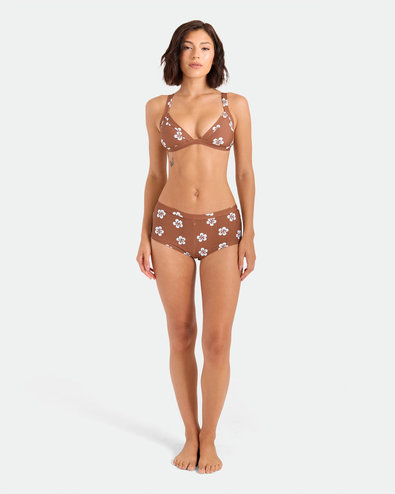 A woman stands barefoot against a white background, wearing Volcom's Women's Coco Retro Bloom Surf Shorts, a brown bikini bottom with white flower patterns made from recycled nylon, showcasing cheeky coverage.