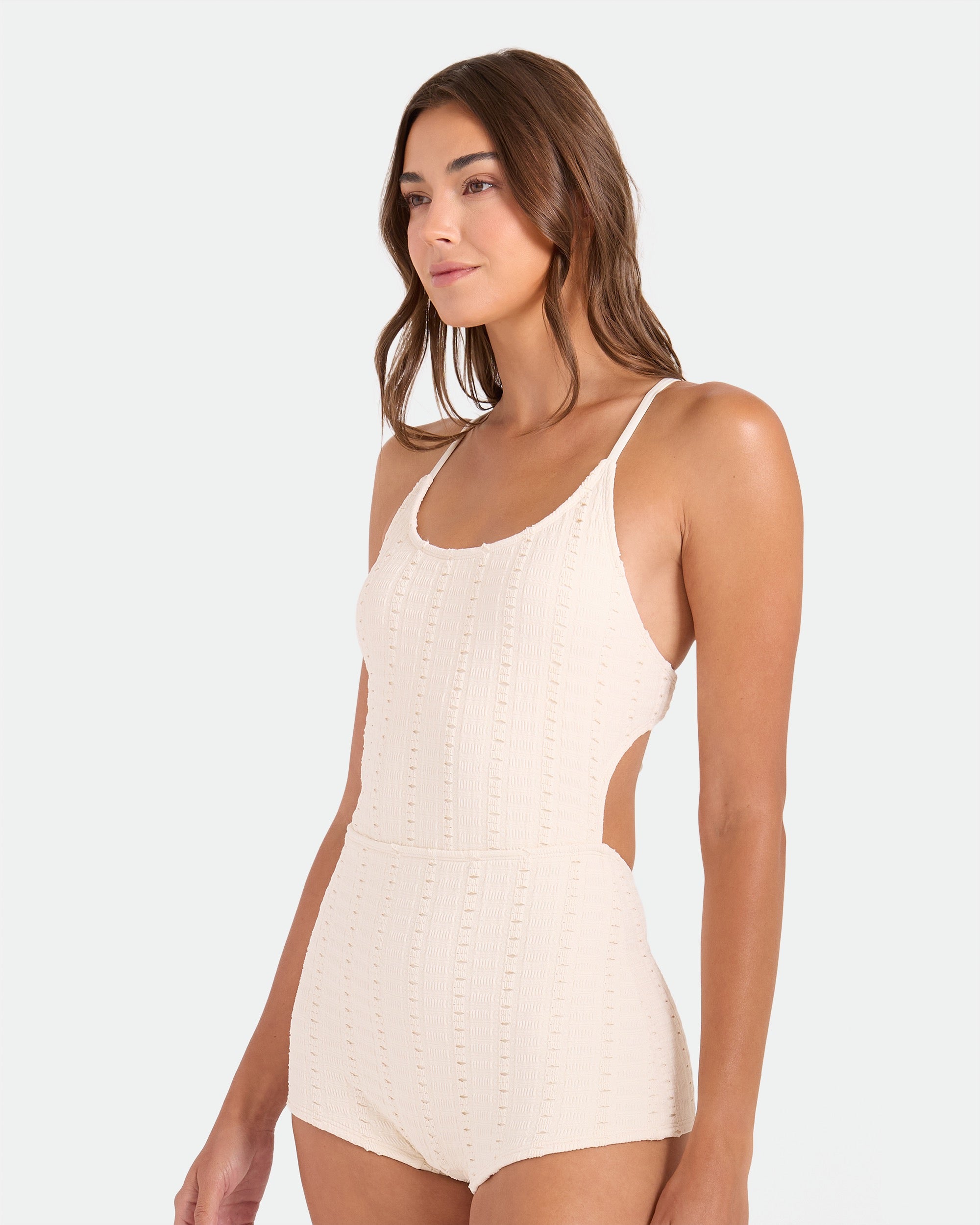 A woman with long brown hair wears the Volcom Women's Coco Low Tide Surf Suit One Piece, a cream textured swimsuit with thin straps and an adjustable tie back, standing against a plain light background.