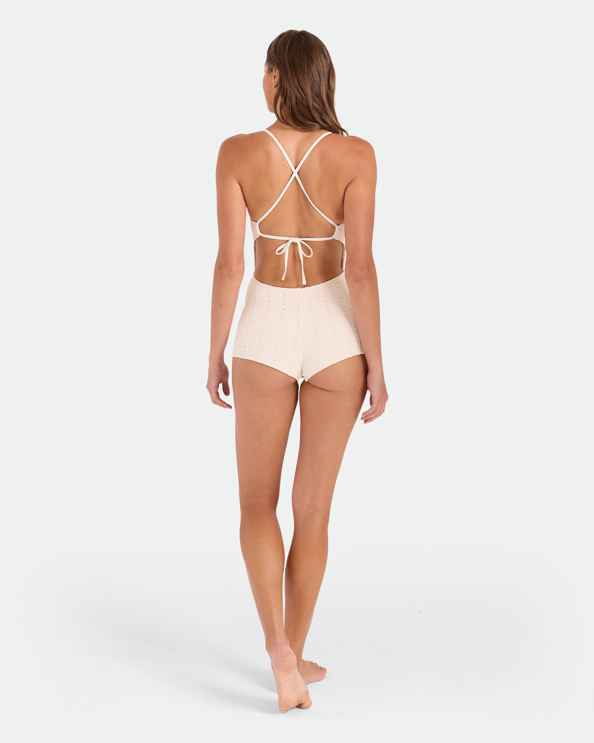 A woman with long brown hair stands barefoot, facing away, wearing the Volcom Women's Coco Low Tide Surf Suit One Piece in cream with a high waist and adjustable tie back straps, set against a plain white background.