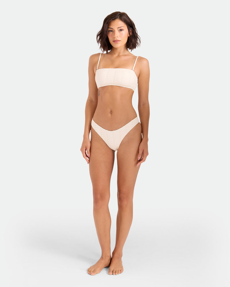 A woman with short brown hair stands barefoot in a Volcom Women's Coco Low Tide Bandeau Bra—a minimalist light beige bikini top with thin, removable, adjustable straps and eyelet details—against a plain light gray background.