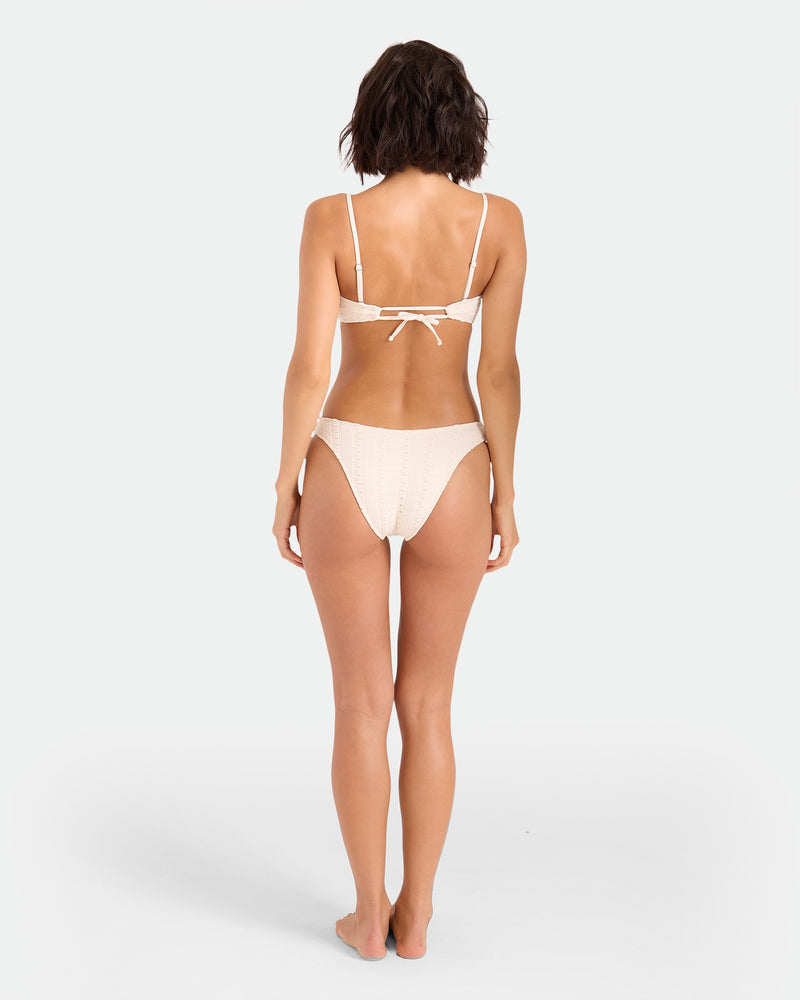 A woman with short brown hair stands barefoot facing away, modeling the Volcom Women's Coco Low Tide Bandeau Bra—minimalist swimwear with removable adjustable straps and a back tie—against a plain white background.