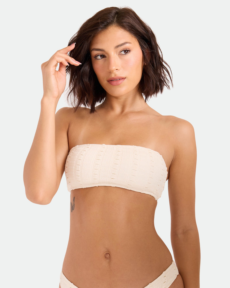 A woman with short dark hair models the Volcom Women's Coco Low Tide Bandeau Bra, a minimalist, textured beige strapless bikini top. She raises one hand near her face and looks slightly to the side against a plain light background.