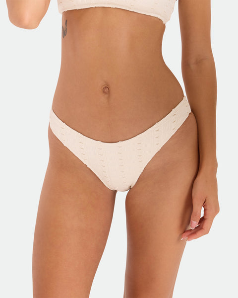 A woman models the Volcom Women's Coco Low Tide 90S V Bottom Skimpy bikini in cream, featuring textured fabric and minimal coverage, posing against a plain white background with a butterfly tattoo on her left side.