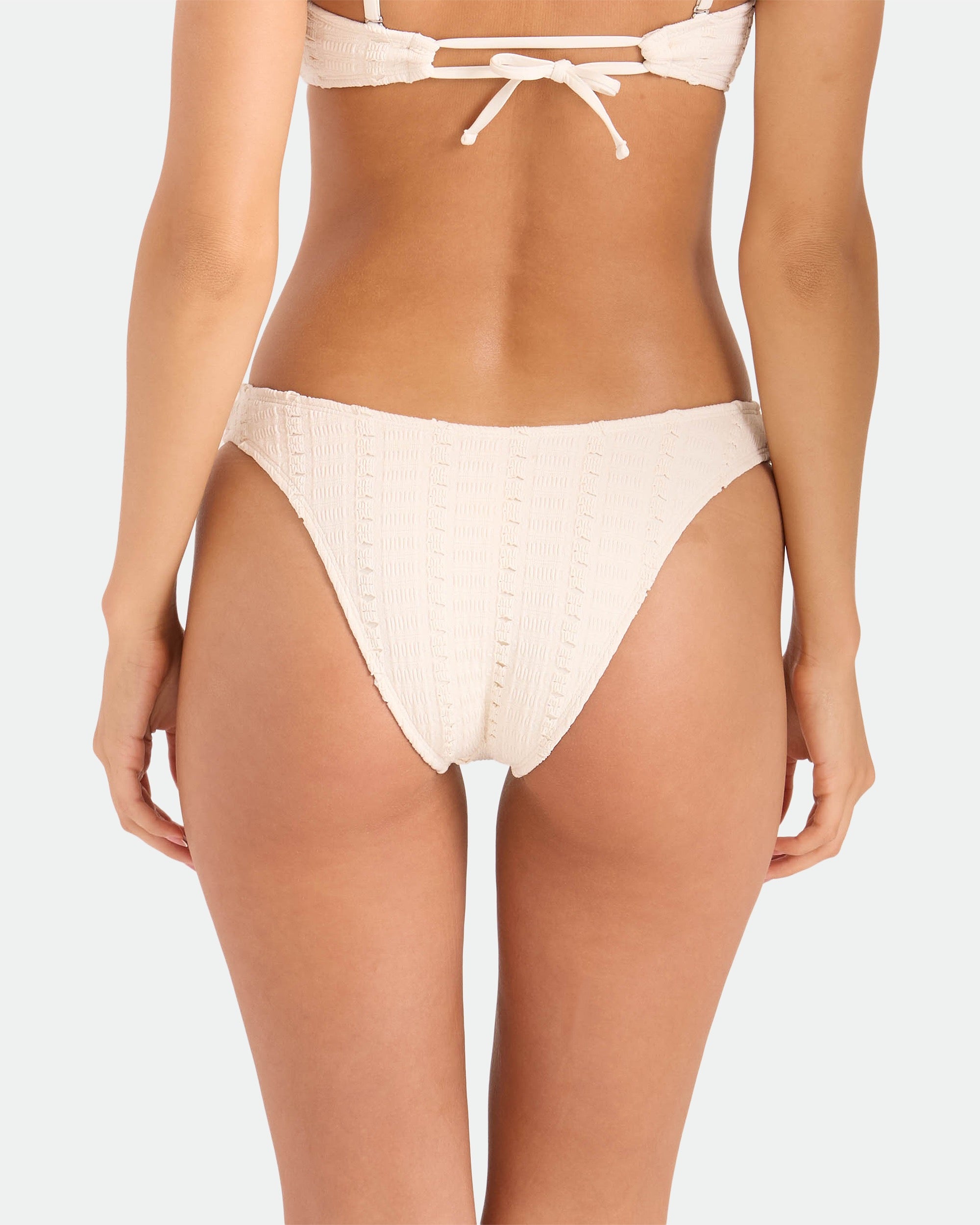 A woman is shown from the waist down wearing the Volcom Women's Coco Low Tide 90s V Bottom Skimpy bikini in cream, with textured fabric and minimal coverage, paired with a matching top, photographed from behind against a plain background.