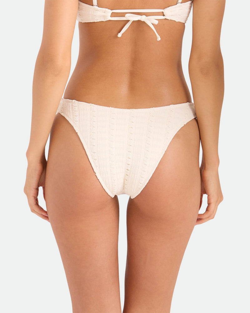 A woman is shown from the waist down wearing the Volcom Women's Coco Low Tide 90s V Bottom Skimpy bikini in cream, with textured fabric and minimal coverage, paired with a matching top, photographed from behind against a plain background.