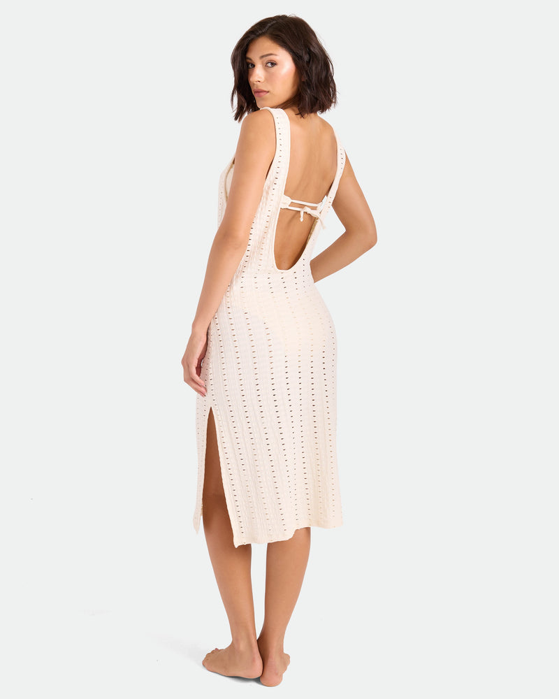 A woman with short dark hair models the Volcom Women's Coco Low Tide Midi Dress, a sleeveless, light-colored crochet tank dress with an open back and side slit, standing barefoot and glancing over her shoulder against a plain backdrop.