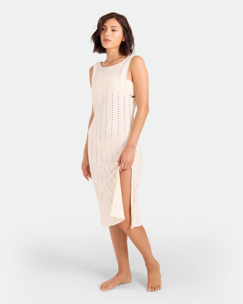 A woman with short brown hair wears the Volcom Women's Coco Low Tide Midi Dress—a sleeveless, light beige, perforated body-con tank dress with side slits—standing barefoot against a plain light background.