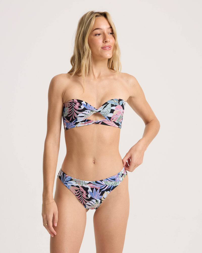 A woman wears the Volcom Women’s Trop Till You Drop Twist Bandeau, featuring a tropical floral print in recycled nylon. She looks to the side with a neutral expression against a plain light background.