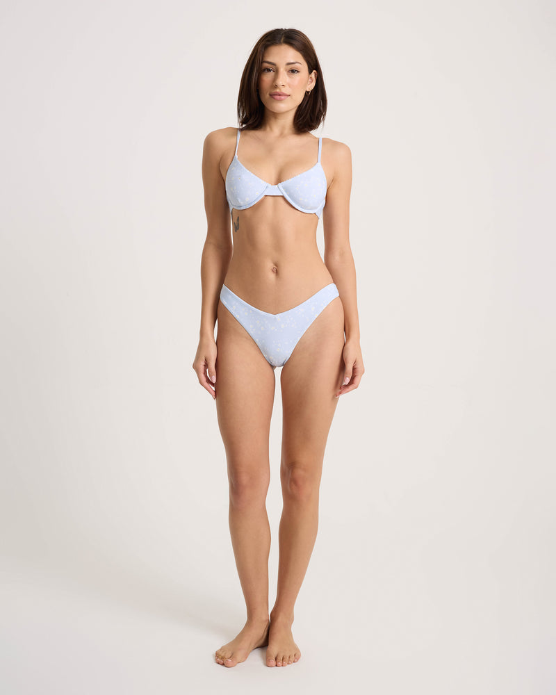 A woman stands barefoot against a plain background, wearing the Volcom Women’s Ditsy Era 90S V Bottom Skimpy in light blue. She faces forward with a neutral expression.