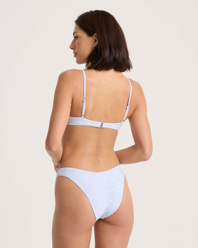 A woman with short dark hair models the Volcom Women’s Ditsy Era 90S V Bottom Skimpy, featuring a light blue floral print, standing with her back to the camera against a plain background.