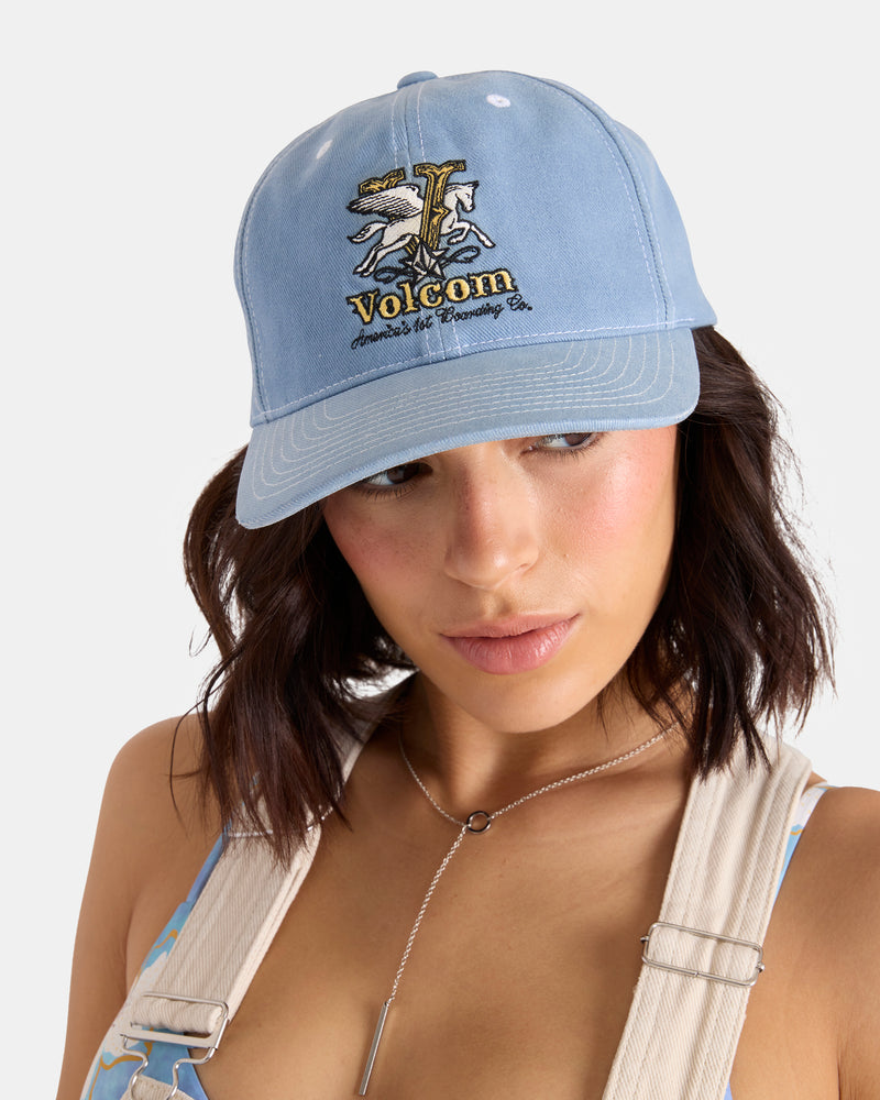 A woman with shoulder-length dark hair wears the Volcom Women's Ameristone Hat, a Horizon Blue denim baseball cap with an embroidered graphic and text, plus beige overall straps and a silver necklace, against a plain background.