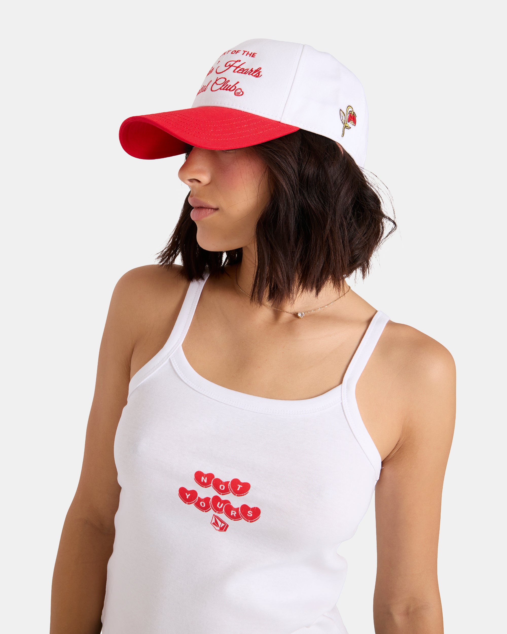 A woman wears the Volcom Women's Breakin Hearts Hat, a white and red cap with embroidered text, a heart detail, and an adjustable strap, paired with a white tank top reading "Hot Young" in red. She looks to the side against a plain background.
