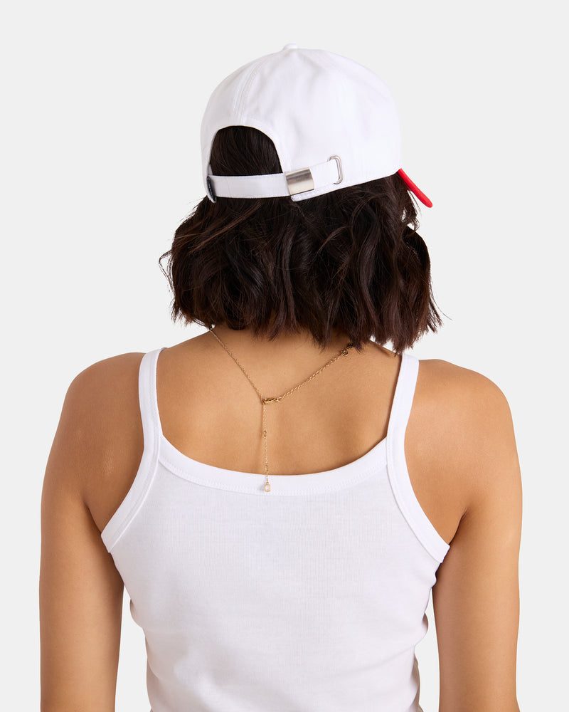 A person with short, wavy dark hair faces away from the camera against a plain background, wearing a white tank top, a delicate gold chain necklace, and the Volcom Women's Breakin Hearts Hat backwards to reveal the adjustable strap.
