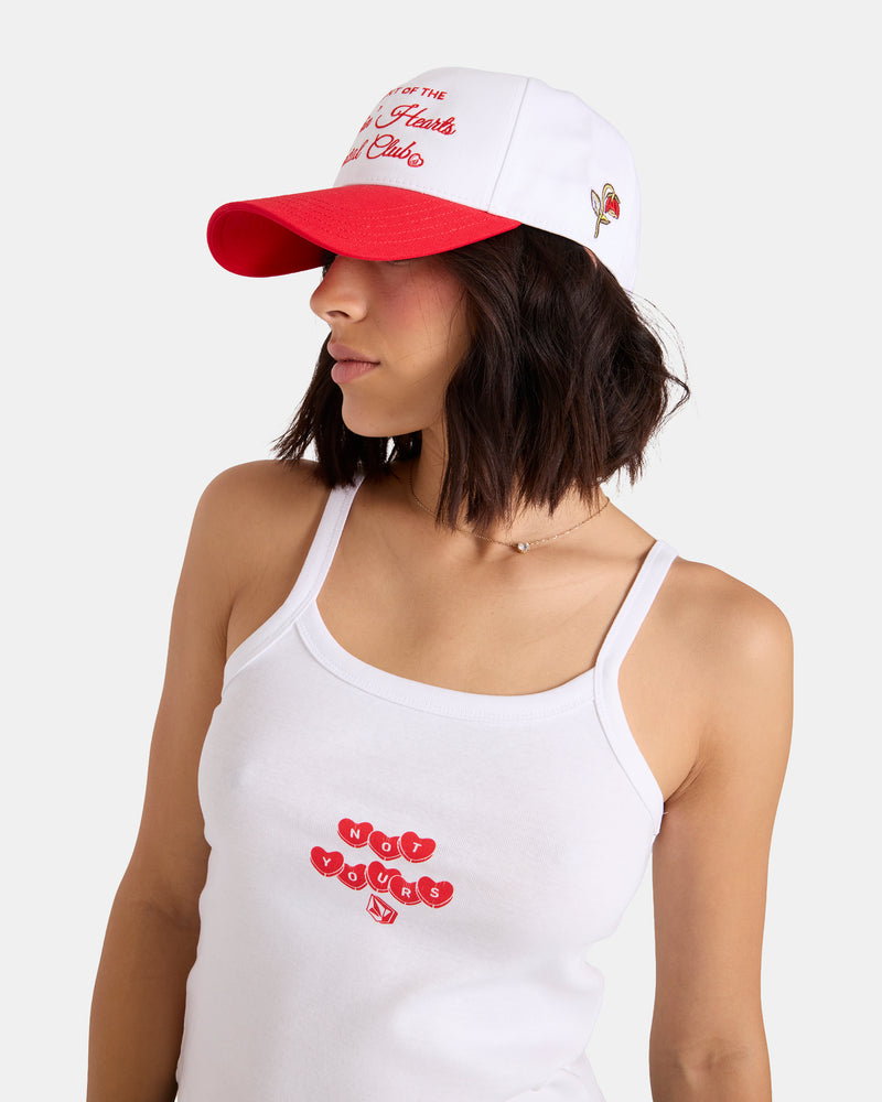 A woman in a white tank top with “HEY YOUNG” in red letters and a Volcom Women's Breakin Hearts Hat, featuring front logo embroidery and a side graphic, looks to her left.