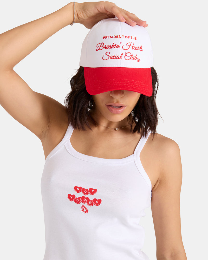 A woman in a white "Not Yours" tank with red hearts wears the Volcom Women's Breakin Hearts Hat—white structured twill with an adjustable back strap—holding the brim and looking down.