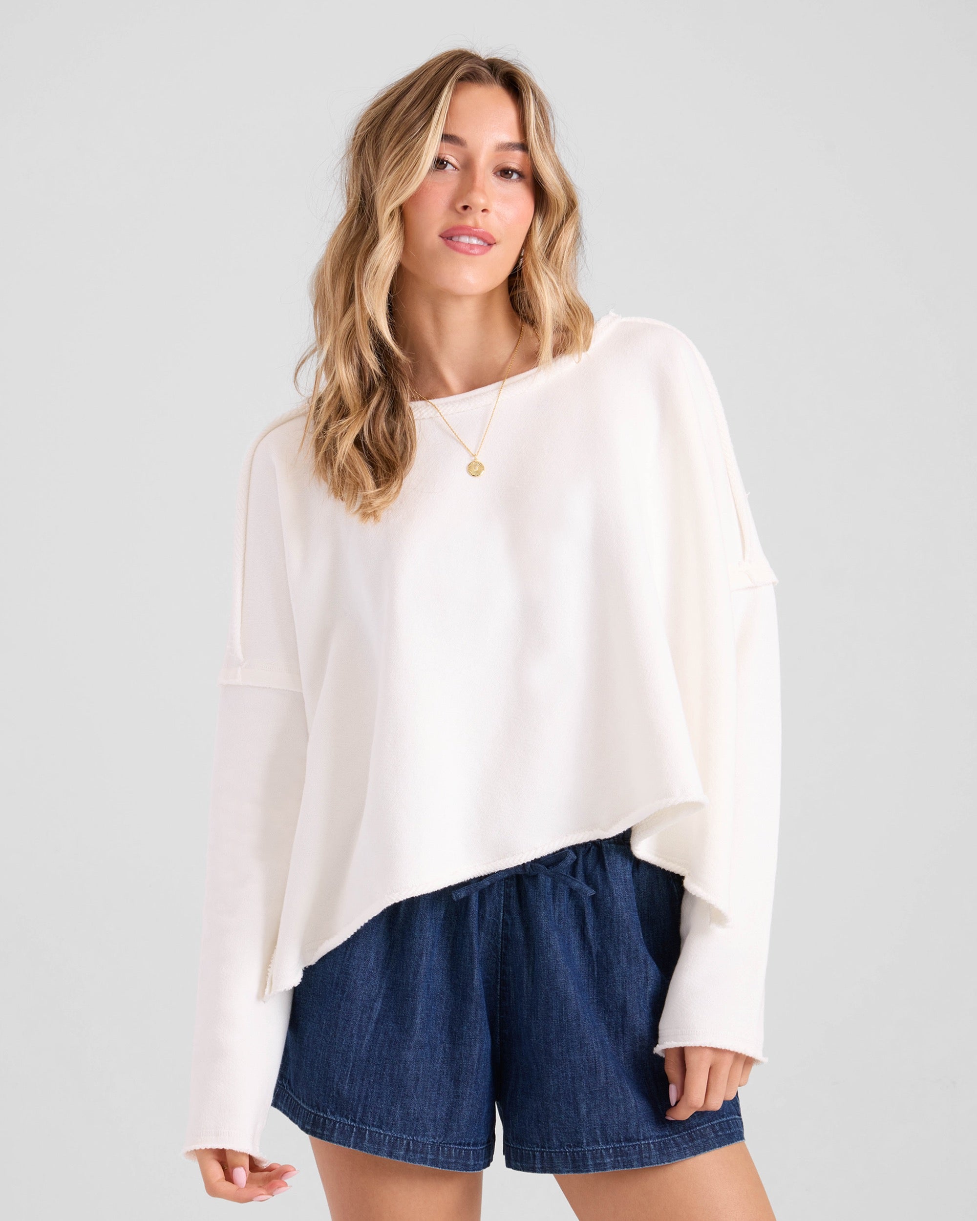 A woman with long, wavy blonde hair wears the Volcom Women's Gnarly relaxed fit sweatshirt and dark blue shorts. The 100% cotton fleece top has a loose, long-sleeve design. She stands against a light grey background, looking relaxed at the camera.