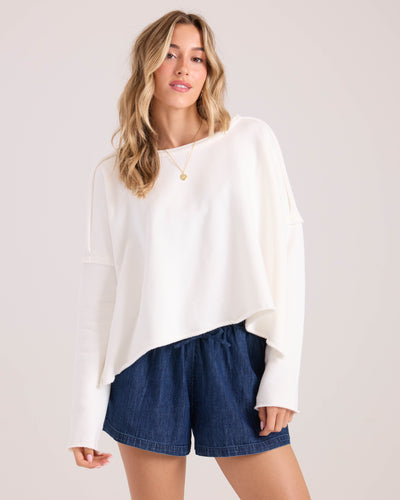 A woman models the Volcom Women's Gnarly, a loose-fitting white long-sleeve top, paired with blue denim shorts and a simple necklace, standing against a plain light background with her hair in loose waves.