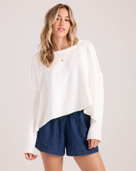 A woman models the Volcom Women's Gnarly, a loose-fitting white long-sleeve top, paired with blue denim shorts and a simple necklace, standing against a plain light background with her hair in loose waves.