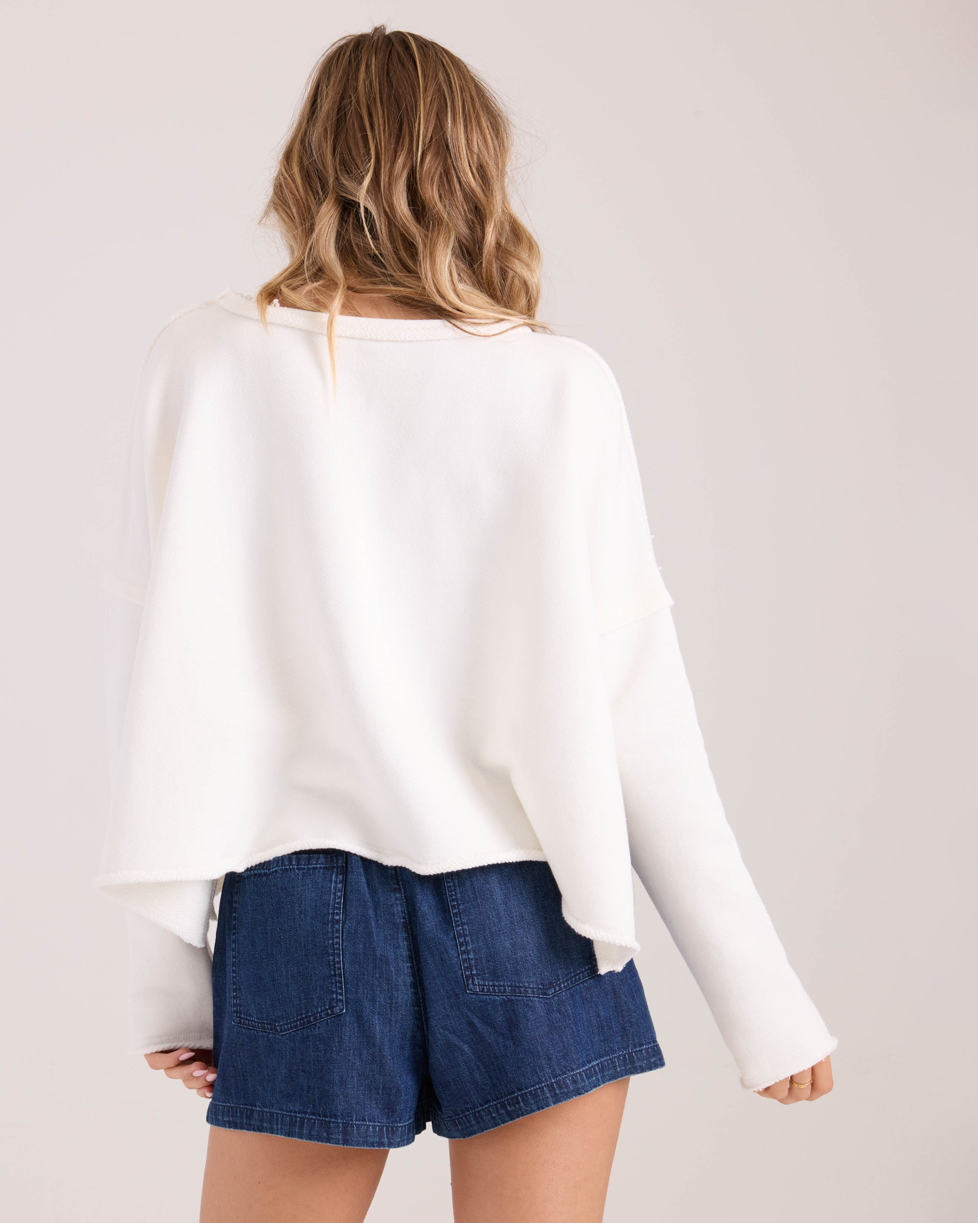 A woman with wavy blonde hair is seen from behind wearing the Volcom Women's Gnarly loose white long-sleeve top and high-waisted blue denim shorts against a plain light background.