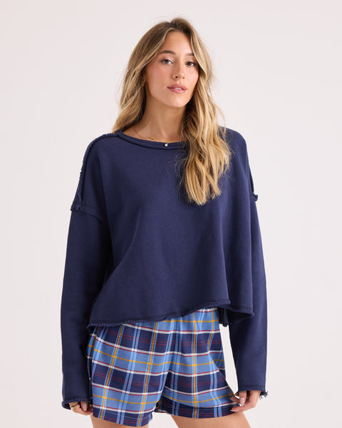 A woman with long blonde hair wears the Volcom Women's Gnarly Sweatshirt in a relaxed fit, paired with blue plaid shorts. She stands against a plain light background, looking forward with a neutral expression.