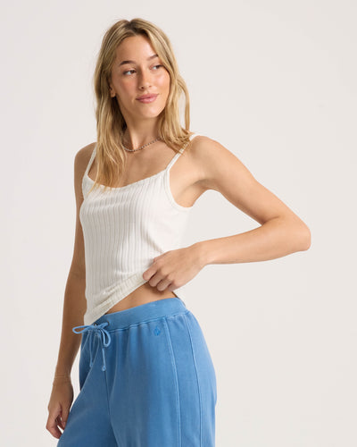 A woman with long blonde hair models the Volcom Women's Lived In Rib Tank and blue drawstring sweatpants, standing against a plain light background while looking to the side and gently pulling at her shirt hem.