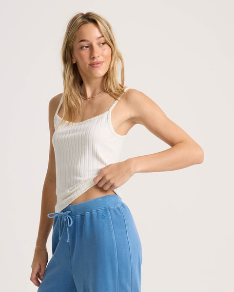 A woman with long blonde hair models the Volcom Women's Lived In Rib Tank and blue drawstring sweatpants, standing against a plain light background while looking to the side and gently pulling at her shirt hem.