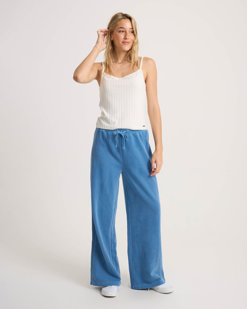 A woman stands against a plain background, smiling and holding her hair with one hand, wearing the Volcom Women’s Lived In Rib Tank in white, paired with loose blue pants and white sneakers.