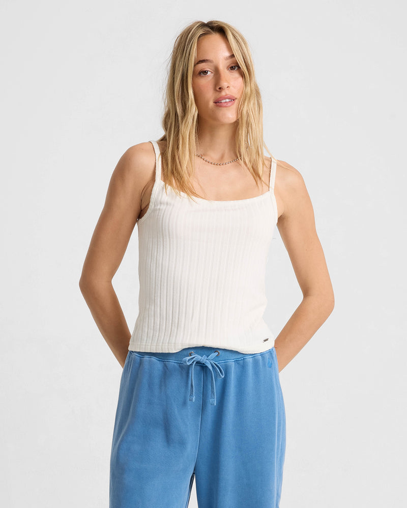 A woman with long blonde hair wears the Volcom Women's Lived In Rib Tank in white, paired with blue drawstring pants, standing against a light gray background and looking at the camera with a relaxed expression.