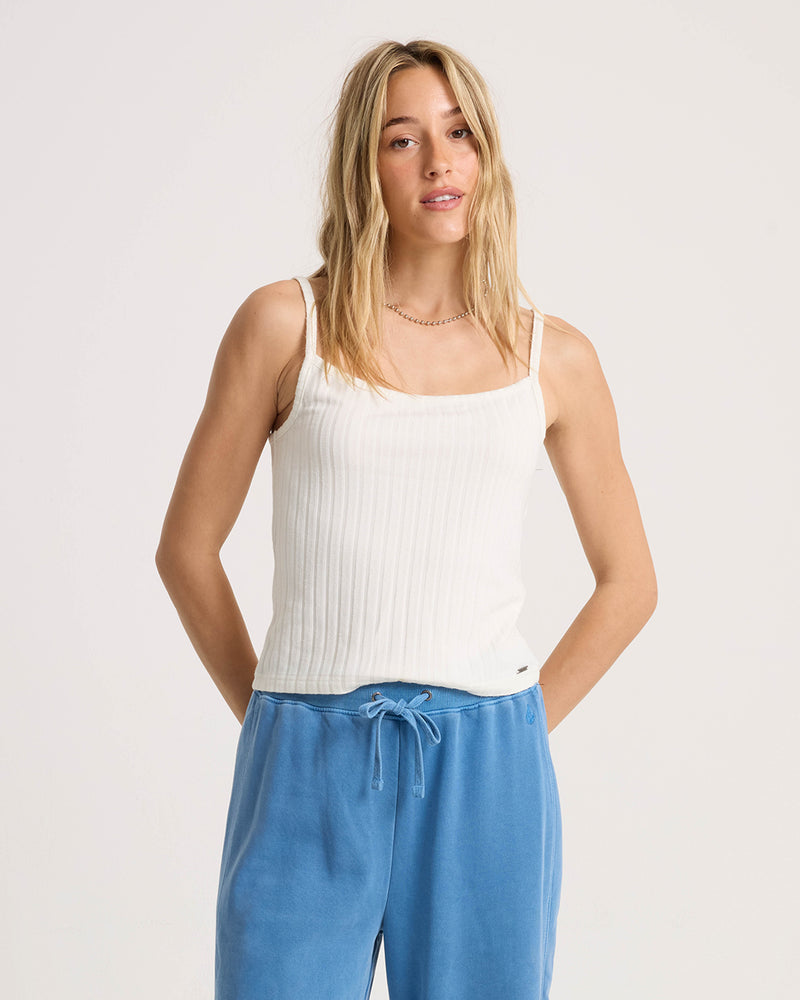 A woman with long blonde hair models the Volcom Women’s Lived In Rib Tank in white, paired with light blue drawstring pants, standing with her hands behind her back against a plain light background.