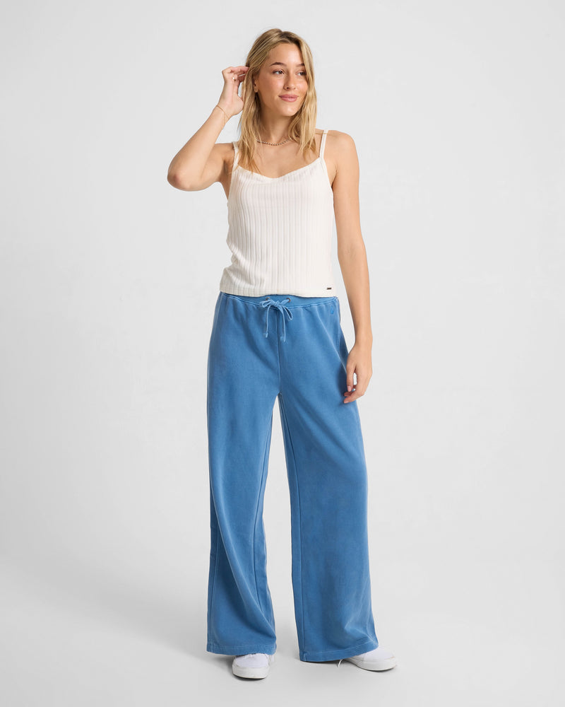 A woman with blonde hair smiles against a white background, wearing the Volcom Women's Lived In Rib Tank, loose blue drawstring pants, and white sneakers. She holds one hand near her head.