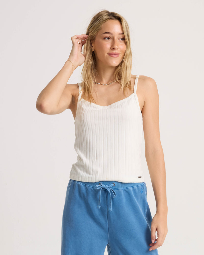 A woman wears the Volcom Women’s Lived In Rib Tank in white with blue drawstring pants, standing against a plain background as she smiles softly and touches her hair.