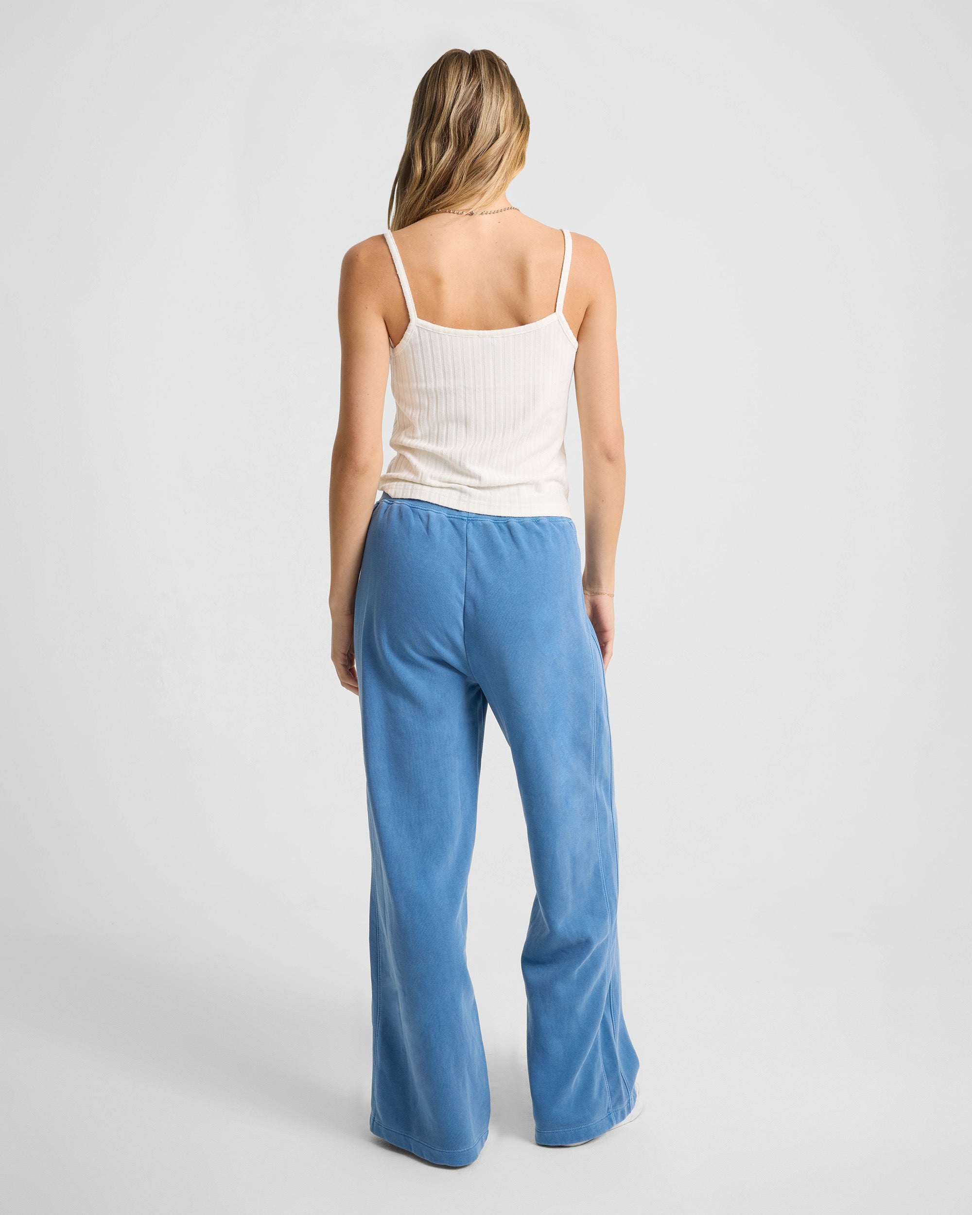 A woman faces away from the camera, wearing the Volcom Women's Lived In Rib Tank in white with spaghetti straps and loose blue pants, set against a plain light backdrop.