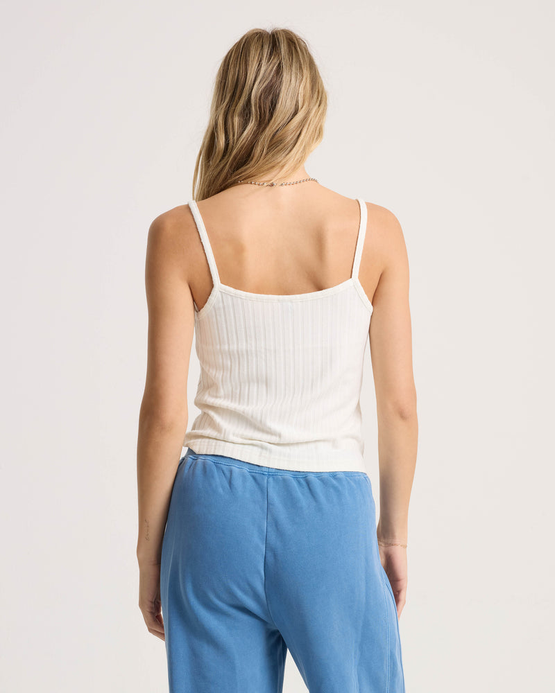 A woman with her blond hair down stands against a plain, light background, wearing the Volcom Women’s Lived In Rib Tank in white and blue pants.