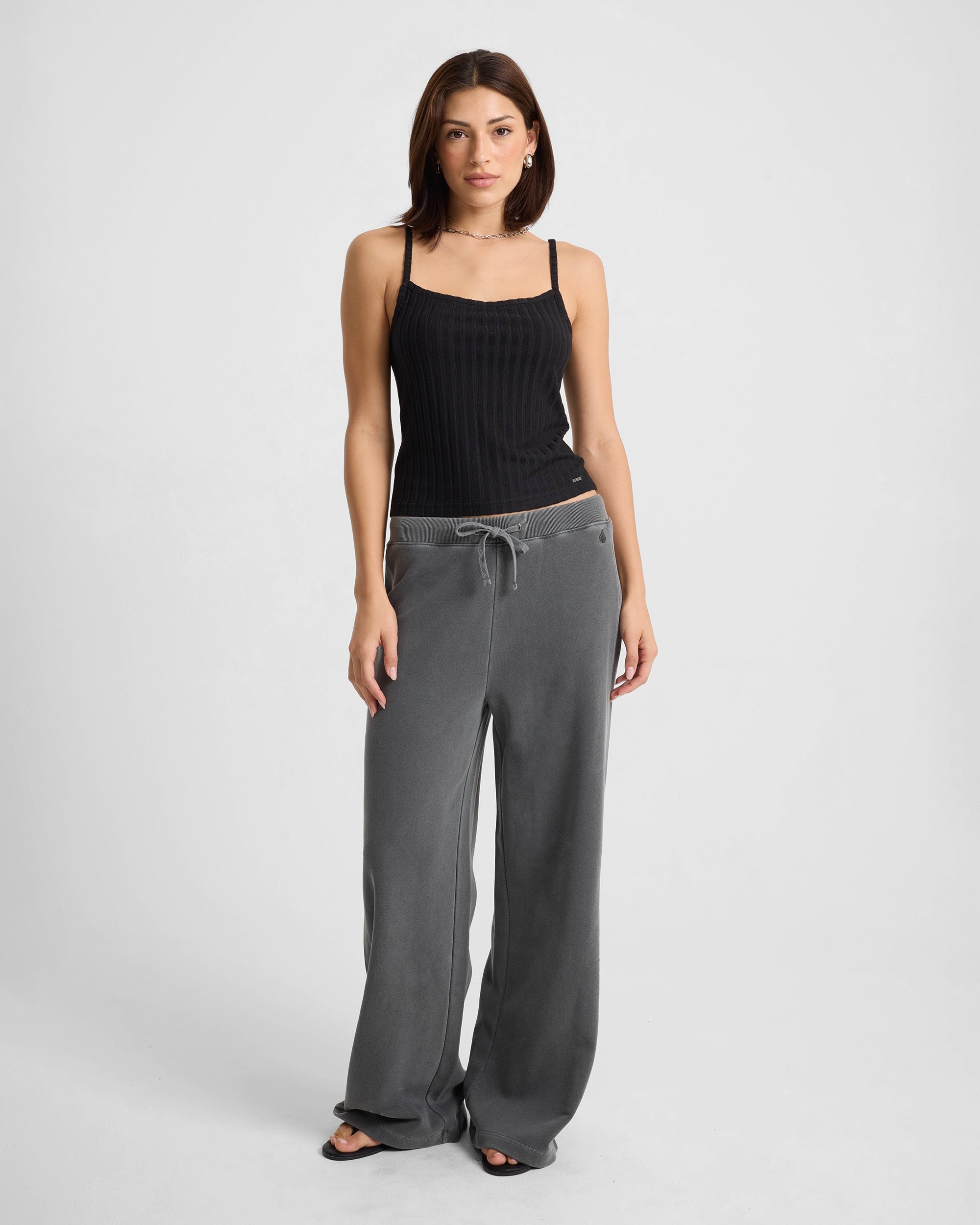 A woman stands facing forward in a Volcom Women's Lived In Rib Tank and loose-fitting gray drawstring pants with black sandals, set against a plain light background.