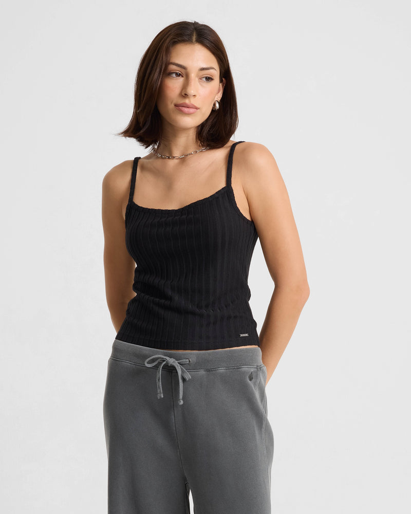 A woman with shoulder-length brown hair wears the Women's Lived In Rib Tank by Volcom in black and gray drawstring sweatpants, standing against a plain light background and looking slightly to the side with a neutral expression.
