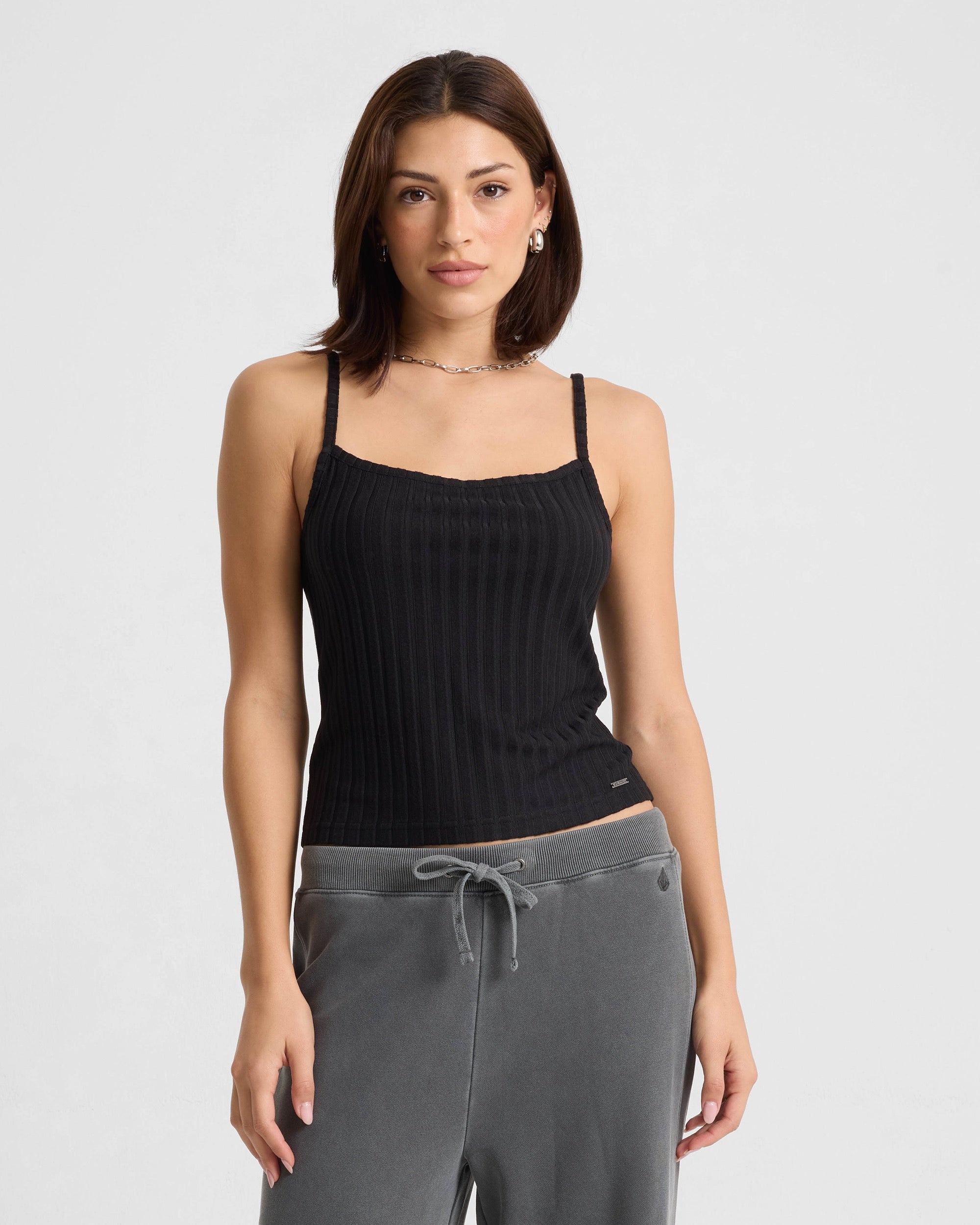 A woman with shoulder-length brown hair wears the Volcom Women's Lived In Rib Tank in black and gray sweatpants, standing against a white background and looking at the camera with a neutral expression.