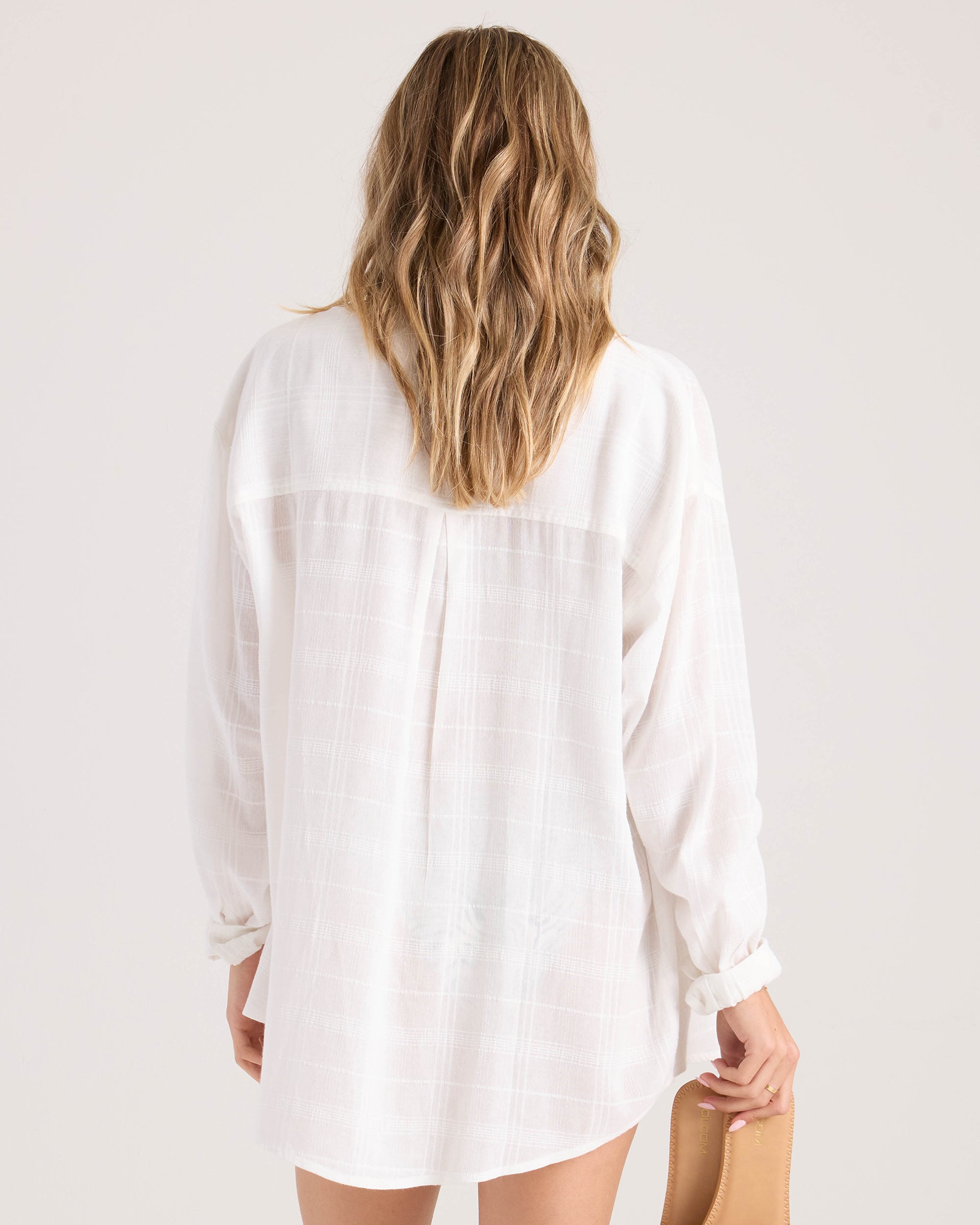 A woman with wavy, light brown hair is seen from behind wearing the Volcom Women's Lived In Button Up 2, a lightweight long-sleeve white shirt, and holding a tan clutch against a neutral background.