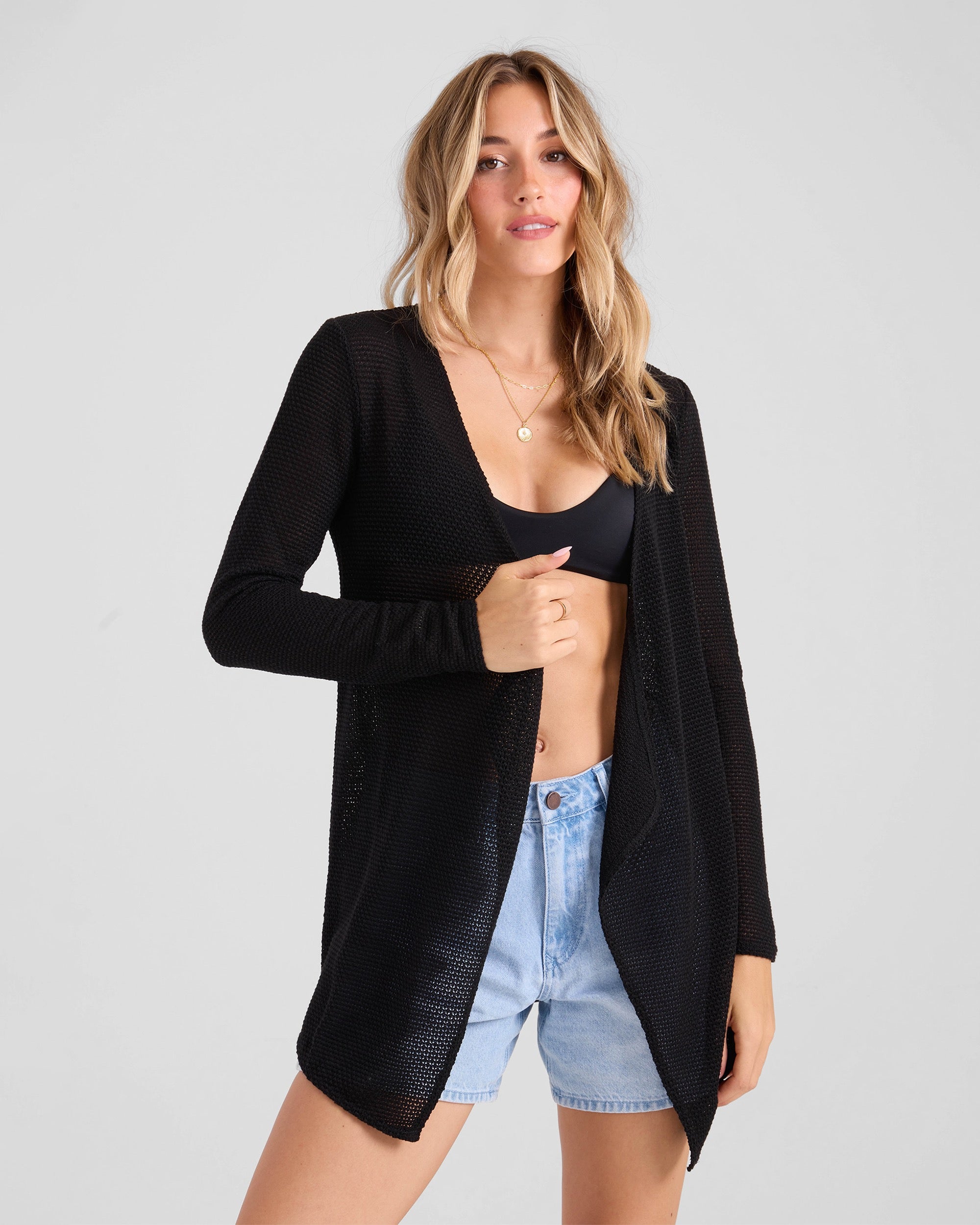 A woman with long, wavy blonde hair wears the Volcom Women's Lived In Go Go Wrap over a black bralette and light blue denim shorts, standing against a plain light gray background.