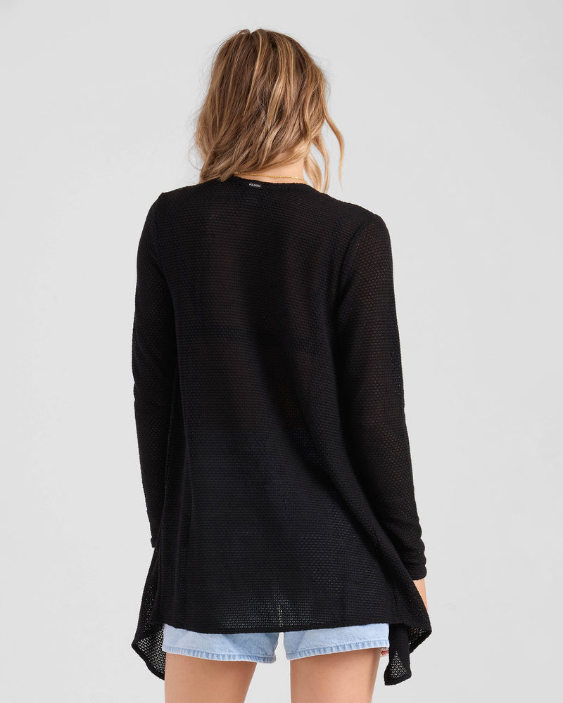 A woman with wavy, light brown hair is seen from the back wearing Volcom's Women's Lived In Go Go Wrap cardigan with light blue denim shorts against a plain light background.