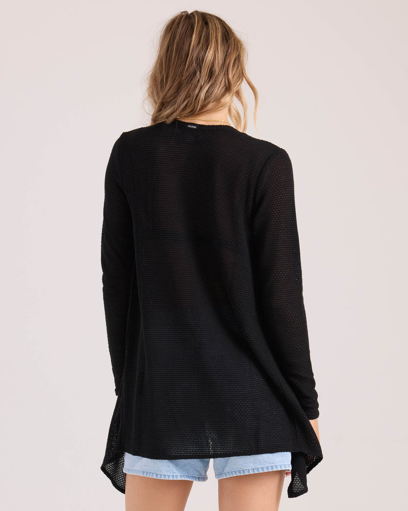 A person with wavy, light brown hair is seen from the back wearing the Volcom Women's Lived In Go Go Wrap cardigan in black and light denim shorts, set against a plain light background.