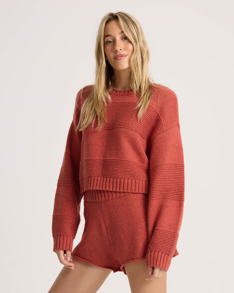 A woman with long blonde hair wears the Volcom Women’s Dayz Like This Sweater—a rust-colored, textured cropped crew sweater with dolman sleeves—paired with matching shorts, standing against a plain white background and looking at the camera with a relaxed expression.